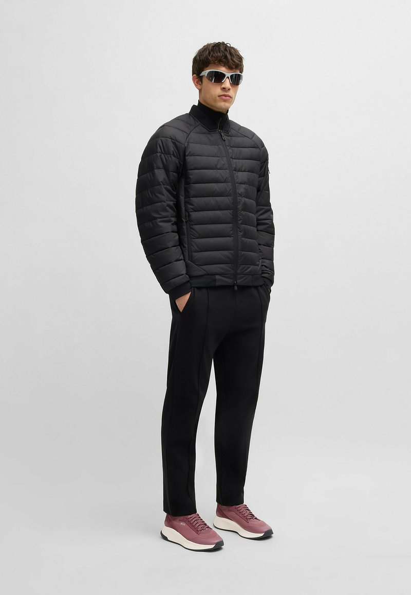 Svart pufferjacka med dragkedja fram och ribbade muddarna, tillsammans med svarta byxor och rosa träningsskor med vita sulor. Solglasögon ingår.