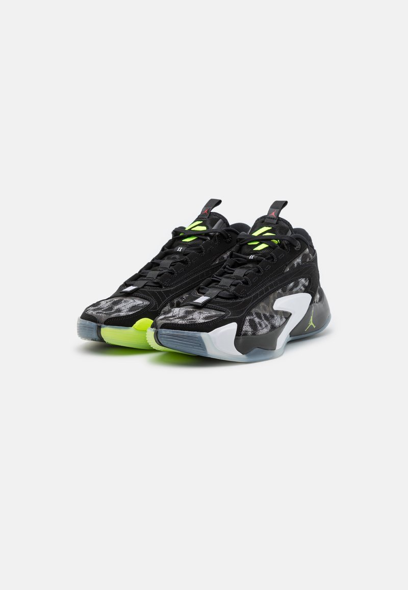 Zapatillas deportivas negras con parte superior de malla, acentos en verde neón y suelas de goma. Presentan formas distintivas y un diseño de tacón transparente.