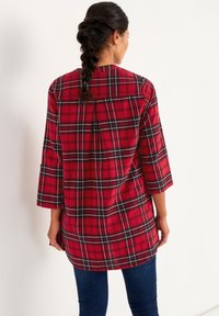Rød ternet tunika med trekvartærmer, lavet af blødt flannelstof. Har sorte og hvide detaljer i ternmønsteret.