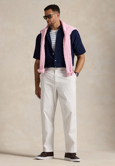 Un homme portant des lunettes de soleil arbore une chemise bleu marine sur un T-shirt rayé, un pantalon blanc et des chaussures marron, avec un pull rose drapé autour des épaules.