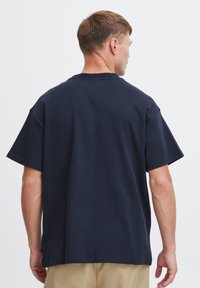 Tricou din bumbac de culoare albastru închis, cu o croială lejeră, mâneci scurte și gât standard în formă de cerc. Textură netedă și design simplu, fără ornamente.
