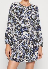 Mekko, jossa on pitkät hihat ja sininen, valkoinen ja vihreä abstrakti kuvio. Kestävä kangas, jossa on röyhelöhelma ja vyötärösidontayksityiskohta.