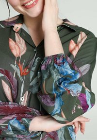 Blusa floreale con sfondo verde scuro, caratterizzata da motivi di foglie colorate in rosa, blu e bordeaux, con dettagli di bottoni e polsini.