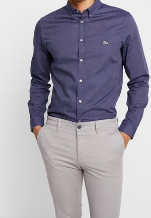 Mann trägt ein eng anliegendes, dunkelblaues Hemd mit Knöpfen und einem kleinen grünen Logo auf der Brust sowie eine hellbeige Hose, die Hände in den Taschen.