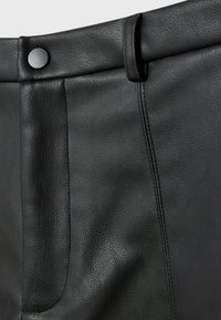 Gros plan sur un pantalon en cuir noir montrant la ceinture, la boucle de ceinture, la couture cousue et une fermeture à bouton noir mat.