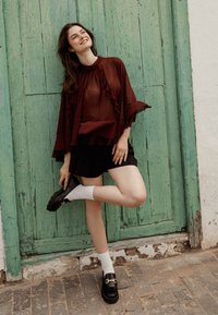 Blusa morada de capas con volantes, pantalones cortos negros y mocasines gruesos negros. Posicionada contra un fondo de madera verde desgastada.