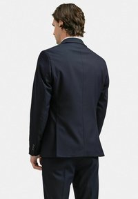 Marinblå kostymjacka sedd bakifrån, med en skräddarsydd passform, slät tyg och en enda slits i mitten. Diskreta stygn på ärmarna.