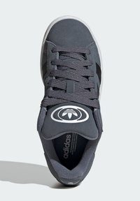 Zapatilla gris con parte superior texturizada, cordones planos y lengüeta acolchada. Presenta rayas negras y un logo blanco en la lengüeta.