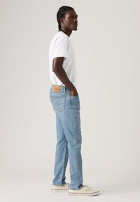 Ανοιχτό μπλε τζιν με ίσια γραμμή, με δερμάτινη ετικέτα μάρκας στη πίσω μέση, συνδυασμένο με απλό λευκό T-shirt και αθλητικά παπούτσια.