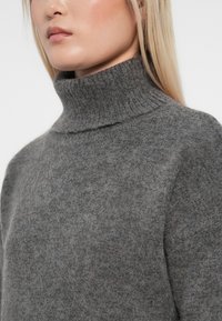 Pulover gri tricotat cu guler înalt, fabricat din lână moale, cu un guler elastic și o croială lejeră, punând în valoare o textură netedă și un design minimalist.