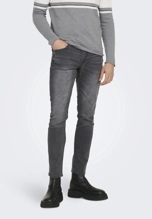 Homme portant un jean slim noir délavé, un pull rayé gris et blanc, et des bottes en cuir noires, debout avec une main dans la poche.