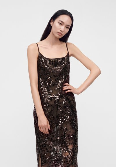 Femme aux longs cheveux noirs portant une robe noire sans manches couverte de sequins brillants, debout une main sur la hanche sur un fond uni.