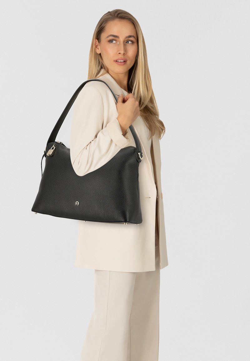 Sac à main en cuir noir avec une surface texturée, une silhouette incurvée et des éléments métalliques dorés, porté sur l'épaule avec une tenue claire.