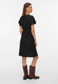 Robe noire mi-longue aux genoux, manches courtes, tissu texturé et taille cintrée, associée à des bottes en cuir marron montant aux genoux.