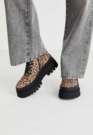 Bottes à lacets en imprimé léopard avec semelles compensées épaisses noires, portées avec un jean gris sur un sol blanc.