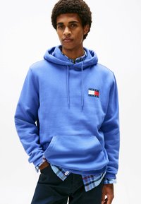Junger Mann, der einen blauen Tommy Jeans Hoodie über einem blauen karierten Hemd trägt, mit den Händen in den Taschen, vor einem weißen Hintergrund stehend.