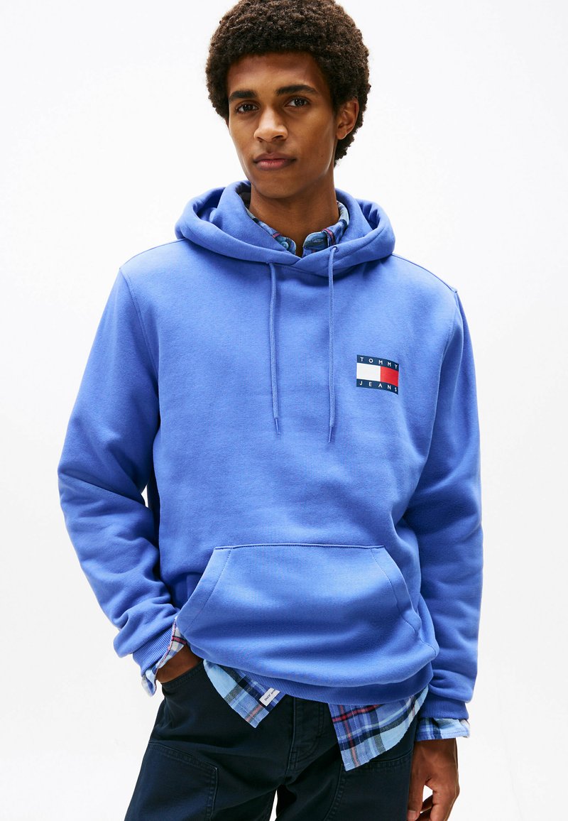 Junger Mann, der einen blauen Tommy Jeans Hoodie über einem blauen karierten Hemd trägt, mit den Händen in den Taschen, vor einem weißen Hintergrund stehend.
