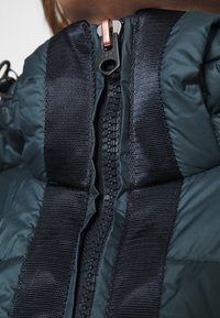 Uppblåsbar jacka med en mörkgrön yta, vadderad textur och en central dragkedja med en metallisk detalj, flankeras av svarta tygpaneler.