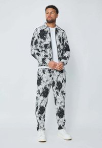 Homme portant une veste et un pantalon marbrés en noir et blanc avec des baskets blanches, se tenant contre un fond uni clair, les mains jointes.