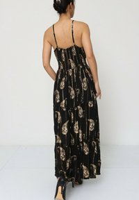 Robe maxi noire avec des bretelles fines, corps froncé et tissu à motif paisley doré. Texture lisse ; longueur jusqu'aux chevilles, accompagnée de talons noirs.