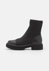 Botín negro hasta el tobillo con suela gruesa, parte superior de tejido elástico y puntera y contrafuerte de cuero, mostrado en perfil lateral sobre fondo blanco.