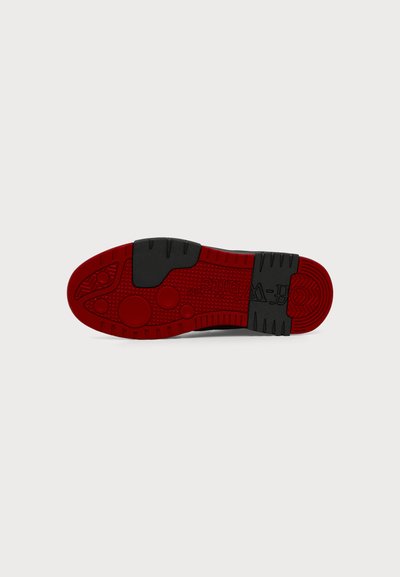 Semelle de chaussure avec une bande de roulement en caoutchouc rouge et noir, présentant des motifs circulaires et des zones de préhension texturées.