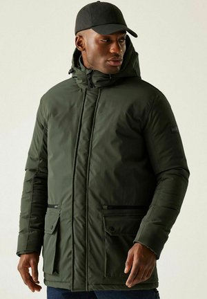 Man die een donkergroene gewatteerde capuchonjas en een zwarte pet draagt, kijkt opzij tegen een effen lichte achtergrond.