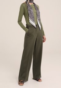 Olive green long-sleeve top paired with wide-leg pants. Black and white patterned silk scarf around the neck. Black sandals with gold accents.