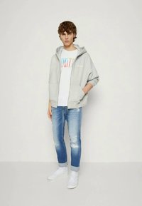Sweatshirt cinza com fecho, sobre uma t-shirt branca com texto multicolorido, combinado com jeans claros esfarrapados e sneakers brancos.