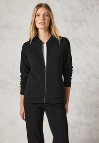 Černá zipová bunda vyrobená z texturovaného materiálu, s límcem ve stylu bomber a žebrovanými manžetami. V kombinaci s bílým tričkem a černými kalhotami.