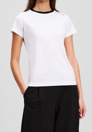 Kvinde iført en kortærmet hvid T-shirt med sort krave og sorte bukser, stående med den ene hånd i lommen foran en ensfarvet baggrund.