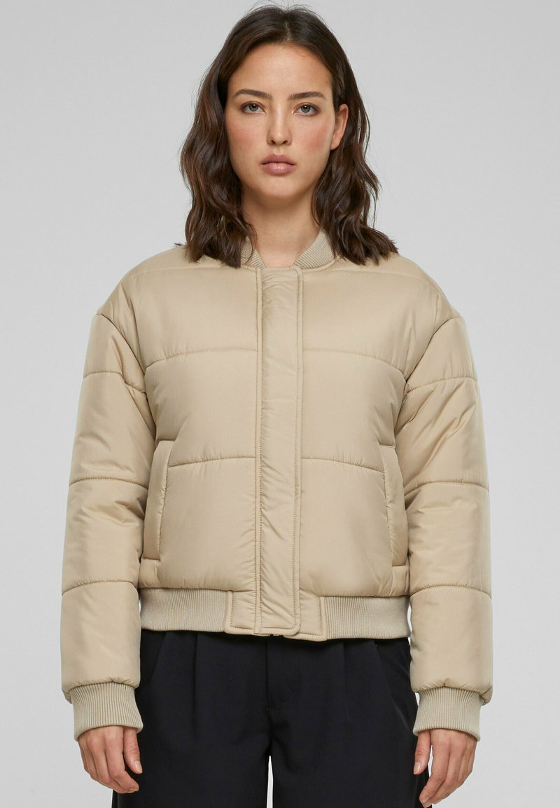 Beige puffjacka med en kortad design, ribbade ärmslut och en dragkedjelös front. Har stickade horisontella paneler för textur och värme.