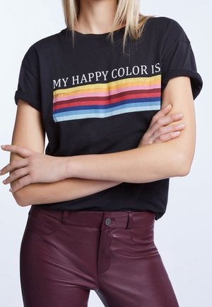 Persona con camiseta negra con rayas horizontales de colores y texto "MY HAPPY COLOR IS", combinada con pantalones de piel rojo oscuro.