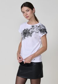 Camiseta blanca de manga corta con un patrón abstracto negro en el frente. La tela es suave y se combina con una minifalda ajustada de cuero negra.
