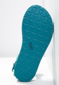 Sole di sandalo blu con un motivo di battistrada triangolare geometrico e il logo "Teva" al centro, posizionata in verticale su uno sfondo bianco.