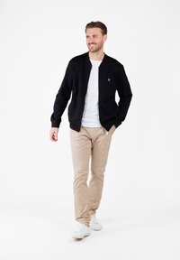 Hombre caminando con chaqueta negra con cremallera, camiseta blanca, pantalones beis y zapatillas blancas sobre un fondo blanco.