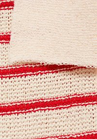 Plaid en crochet rayé crème et rouge avec un tissu tricoté crème plié dessus, montrant des motifs de fil texturés.