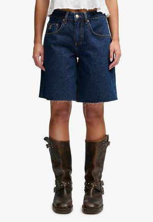 Femme portant un short en jean bleu foncé et des bottes en cuir marron mi-mollet avec des détails de boucle et de clous, debout devant un fond blanc.