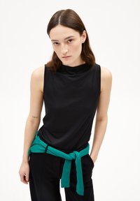 Maglietta nera senza maniche con colletto alto, abbinata a pantaloni neri e una cravatta turchese annodata in vita. Tessuto dalla texture liscia.