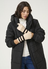 Geacă puffer neagră cu fermoar în față și glugă supradimensionată, asortată cu mănuși crem cu dungi negre. Pulover alb cu guler înalt pe dedesubt.