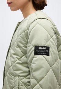 Άτομο που φοράει ανοιχτοπράσινο μπουφάν με επένδυση και ετικέτα της μάρκας ECOALF στο πάνω μέρος του μανικιού, προωθώντας το μήνυμα της βιωσιμότητας.