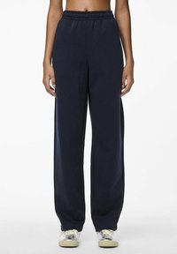 Marineblauwe sweatpants met een elastische tailleband, wijde pijpen en een zachte textuur. Draagt met lichtgekleurde sneakers.