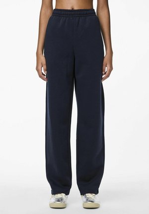 Marineblauwe sweatpants met een elastische tailleband, wijde pijpen en een zachte textuur. Draagt met lichtgekleurde sneakers.