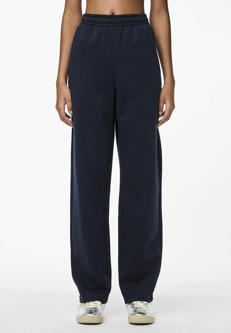 Marineblauwe sweatpants met een elastische tailleband, wijde pijpen en een zachte textuur. Draagt met lichtgekleurde sneakers.