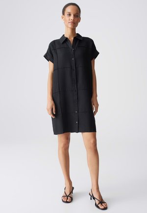 Donna che indossa un vestito camicia nero a maniche corte con bottoni e sandali neri con tacco e cinturini, in piedi su uno sfondo chiaro.