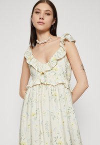 Robe à motif floral en crème, avec un col et des bretelles à volants. Confectionnée en tissu doux et texturé avec des fleurs pastel jaunes et bleues.