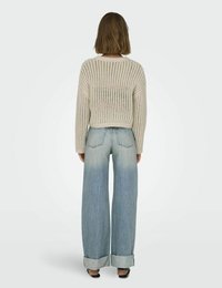 Cremfarbener gestrickter Pullover mit weitem, offenem Maschenwerk und gerippten Bündchen, kombiniert mit hellblauen, locker sitzenden Jeans mit umgeschlagenen Säumen. Rückansicht.