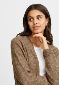 Cardigan lavorato a maglia marrone con texture e polsini a coste, abbinato a un top bianco. La modella ha i capelli scuri di media lunghezza e tiene il mento appoggiato sulla mano.