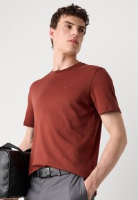 Tricou de culoare ruginită, cu gât rotund, din bumbac, cu mâneci scurte și un logo subtil pe piept. Purtați-l cu pantaloni gri și o geantă neagră.