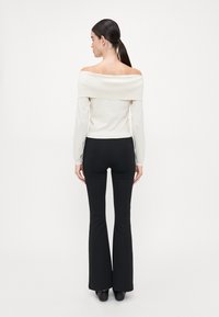 Off-shoulder kremtopp med lange ermer og ribbestruktur, kombinert med svarte flared bukser, vist fra baksiden.